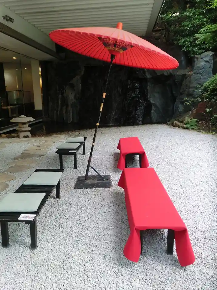 A serene zen garden with white gravel, black stone benches with light cushions, and a large red parasol. Two long benches…