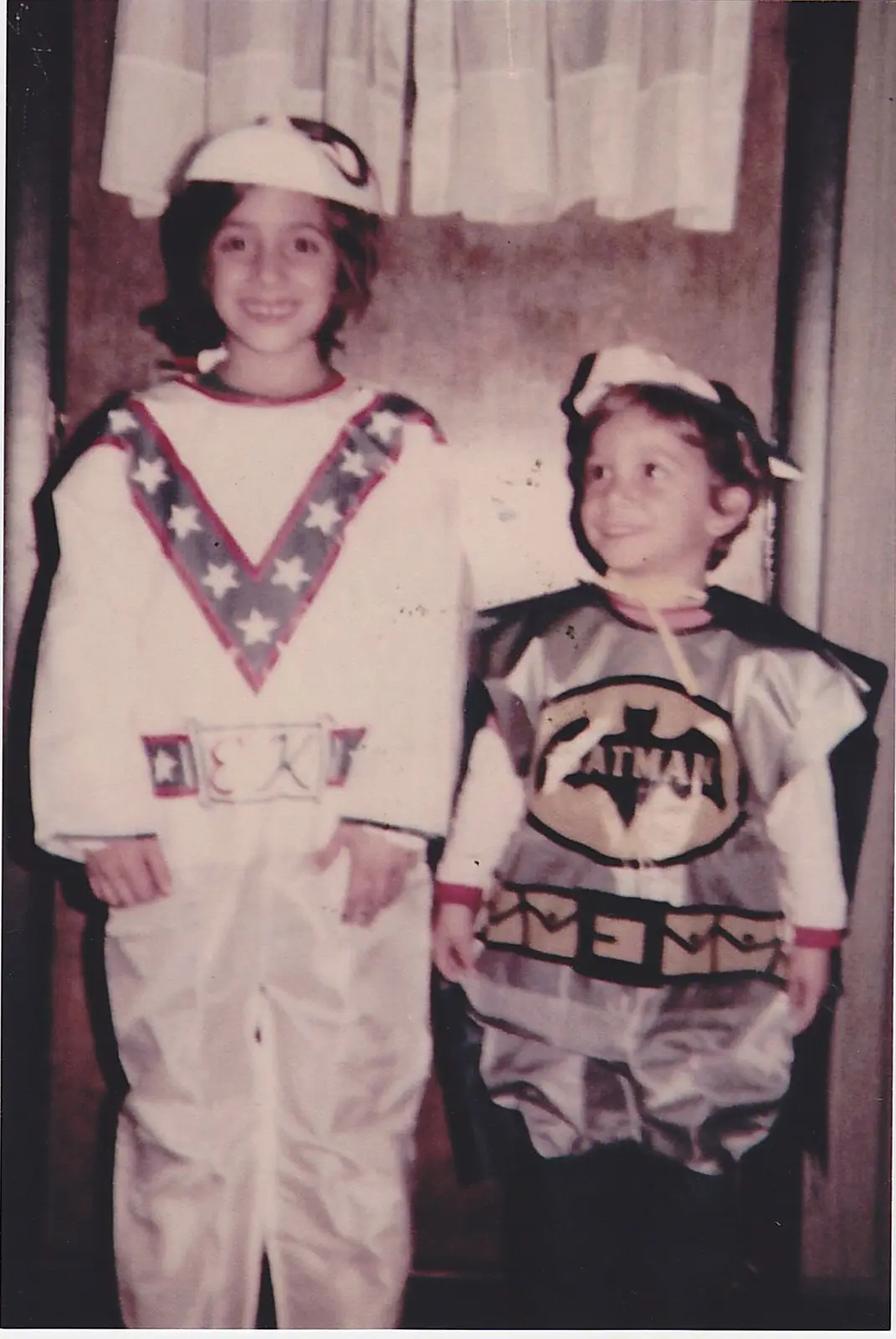 Two young children in vintage superhero costumes pose for a photo; the taller child wears a white costume with a…