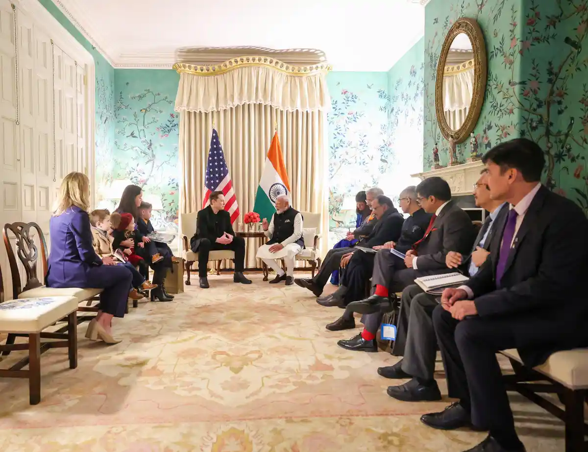 In a formal diplomatic room with floral wallpaper and flags of the United States and India, a man in a black suit sits…