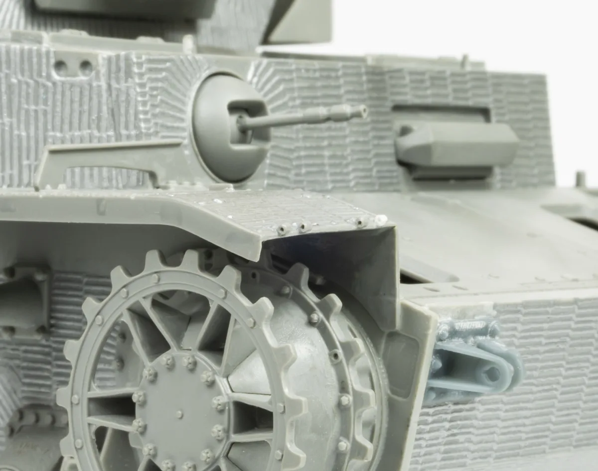 A close-up photo of a grey plastic model tank, showing detailed treads, a gun barrel, and textured armor. The overlaid text…