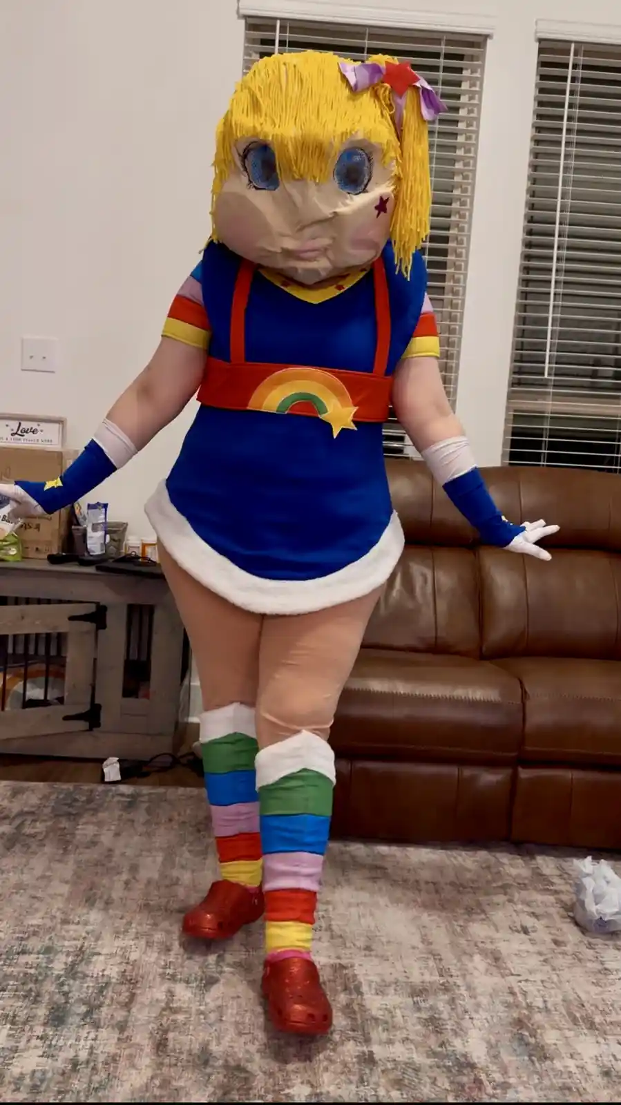A person in a full-body costume of the character Rainbow Brite stands in a living room, wearing a blue dress with rainbow…