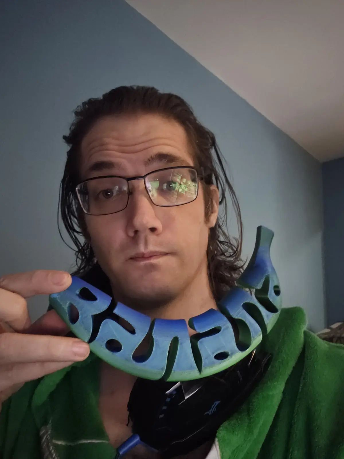 A person with glasses and long hair holds a blue and green 3D-printed necklace shaped like the word 'BANANA' in front of…