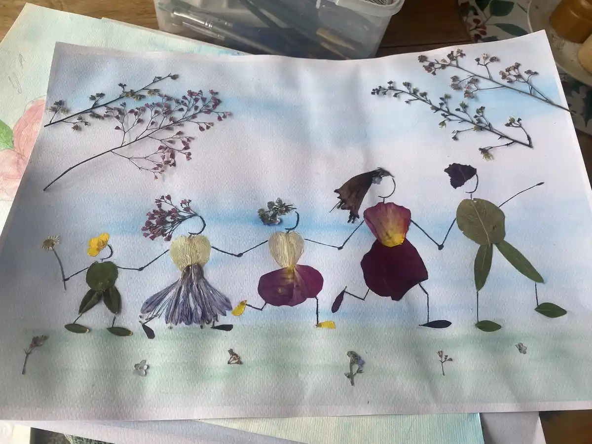 A handmade artwork on watercolor paper depicts five stick-figure people holding hands, each crafted from different dried…