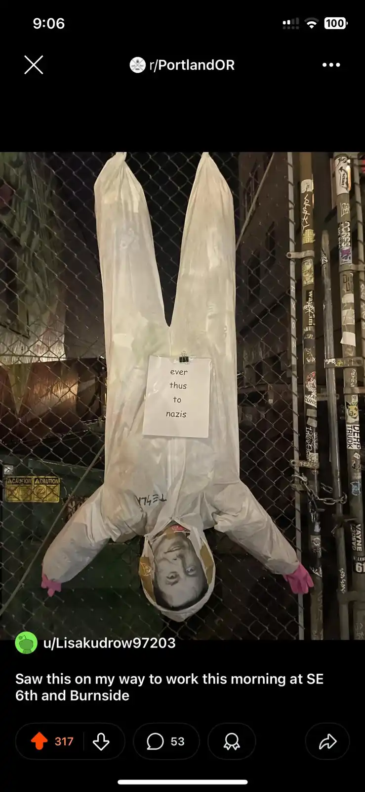 A person in a white hazmat suit with pink gloves is hanging upside down from a chain-link fence at night, with a printed…