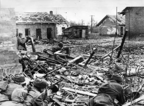 A black-and-white historical photo shows soldiers in a war-torn village with rubble and damaged buildings. Several soldiers…
