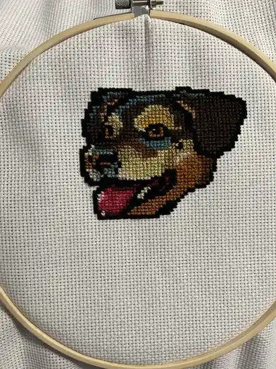 A close-up photo shows a partially completed cross-stitch portrait of a dog's face on white fabric, held taut in a wooden…