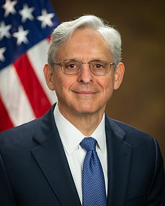 A formal portrait of a man with white hair and glasses, wearing a dark suit and blue tie, standing in front of an American…