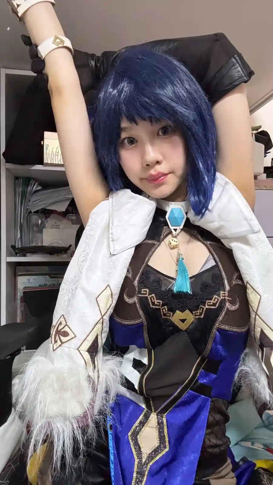 A person in a detailed cosplay outfit with blue hair and white fur-trimmed sleeves is sitting with both arms raised…