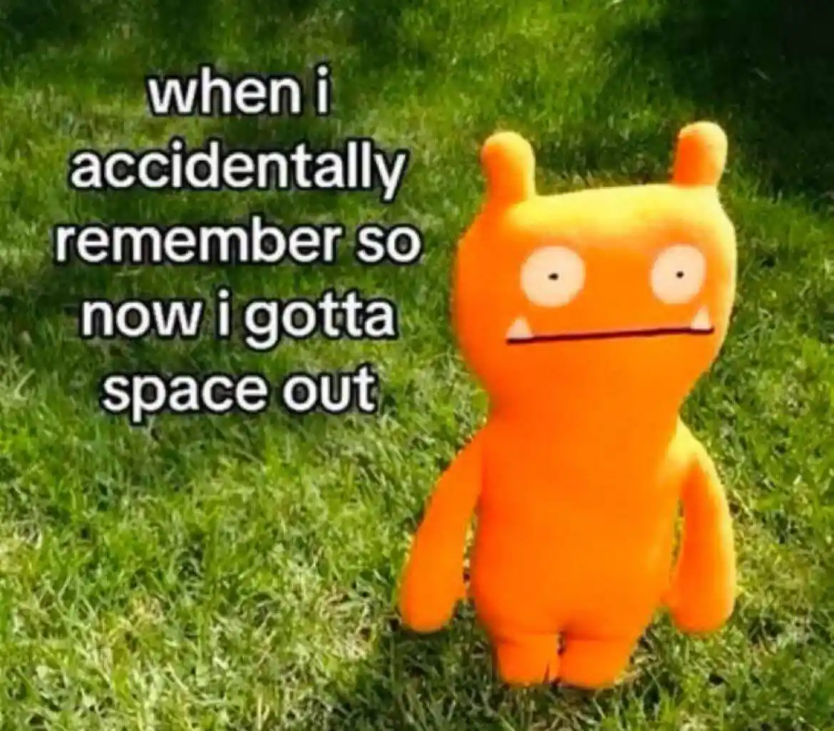 An orange Uglydoll plush toy with a blank, unblinking expression stands on green grass. White text with a black outline is…