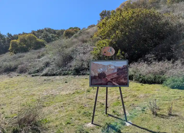 An outdoor interpretive sign on a metal easel stands in a grassy field with hills and trees in the background. The sign…