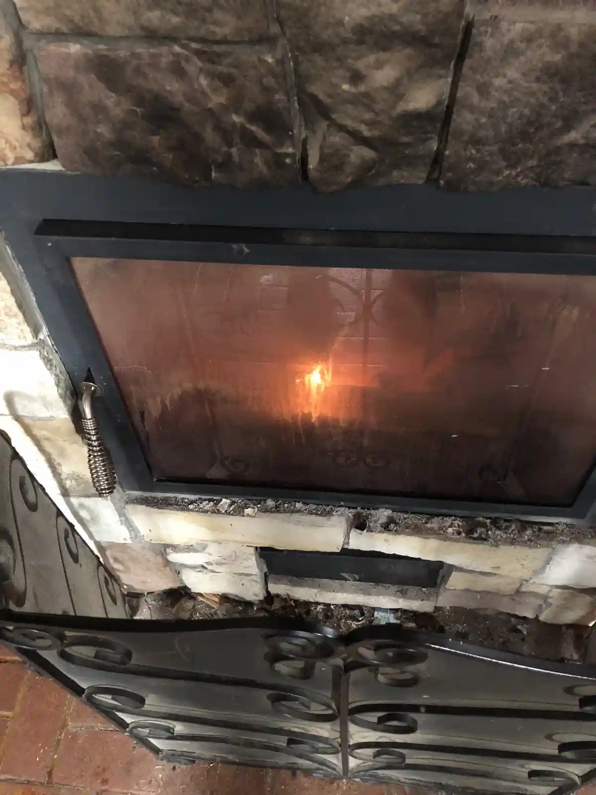 A close-up photo of a stone fireplace with a black metal frame and glass door, showing a small, flickering orange flame…