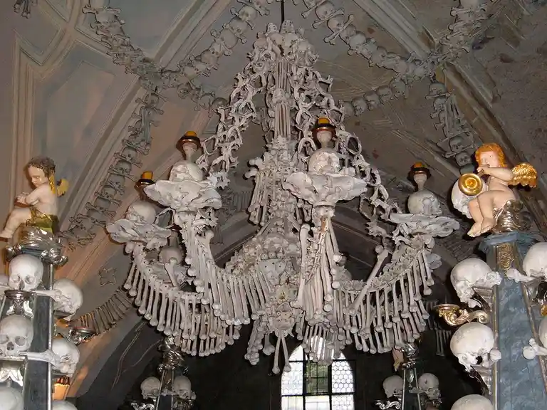 the image shows an ornate chandelier constructed entirely from human bones and skulls, hanging from a vaulted ceiling also…