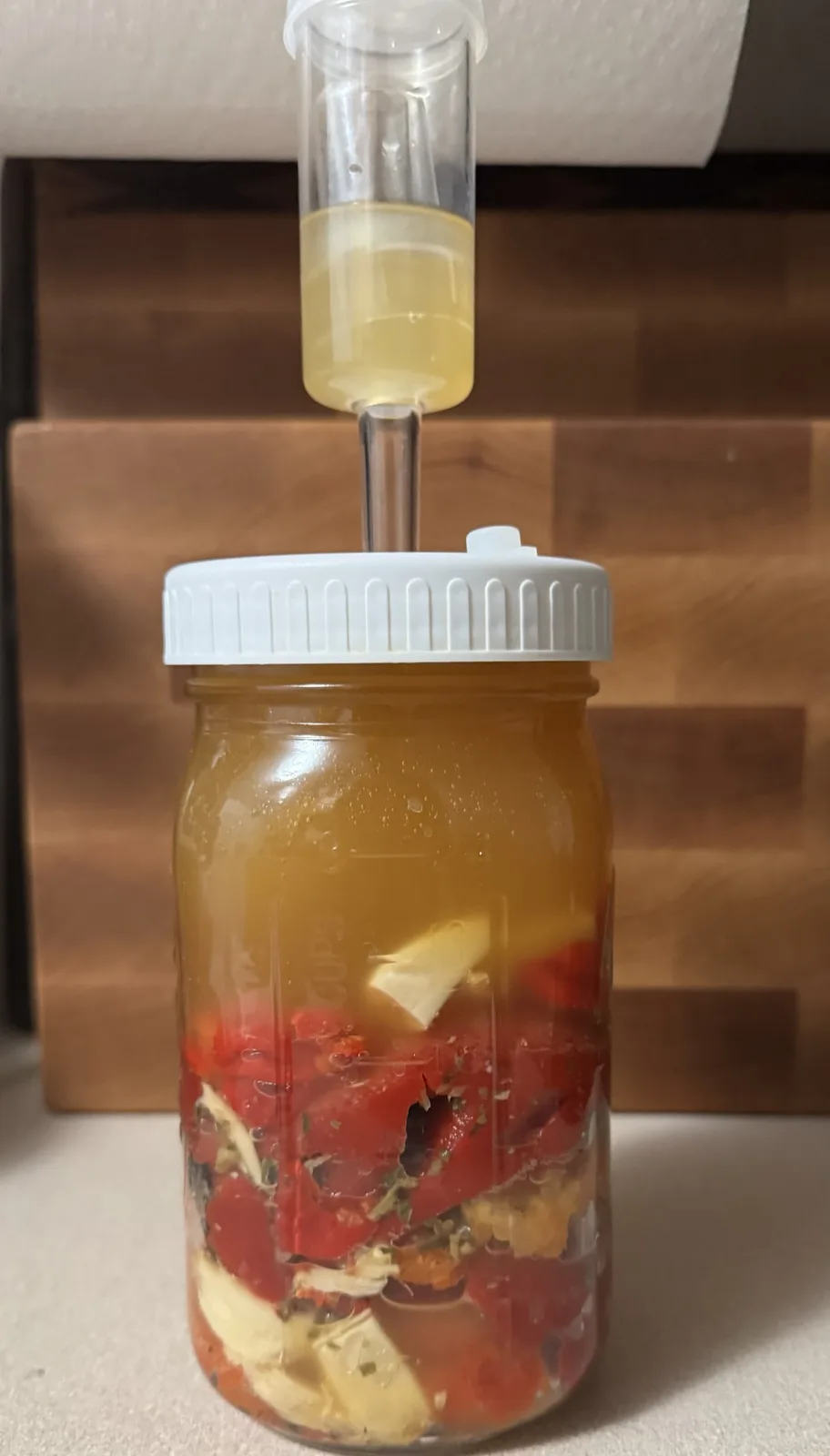 A glass mason jar filled with chopped red vegetables and herbs submerged in brine, sealed with a white lid and a clear tube…
