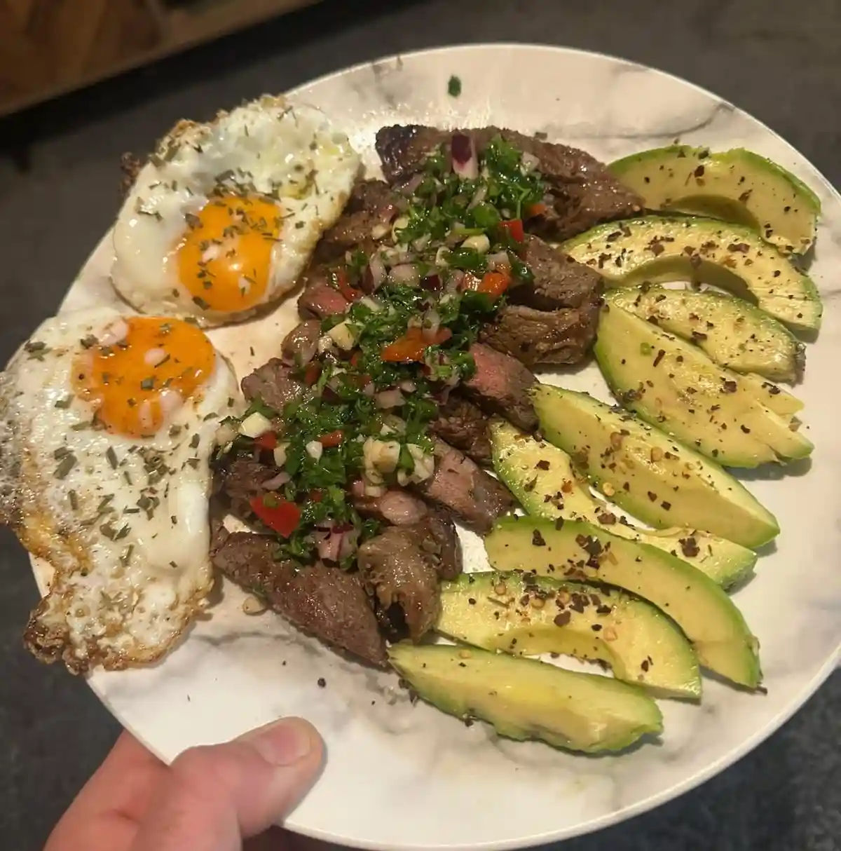 A close-up photo shows a hand holding a white marble-patterned plate with a meal: two sunny-side-up eggs with runny yolks…