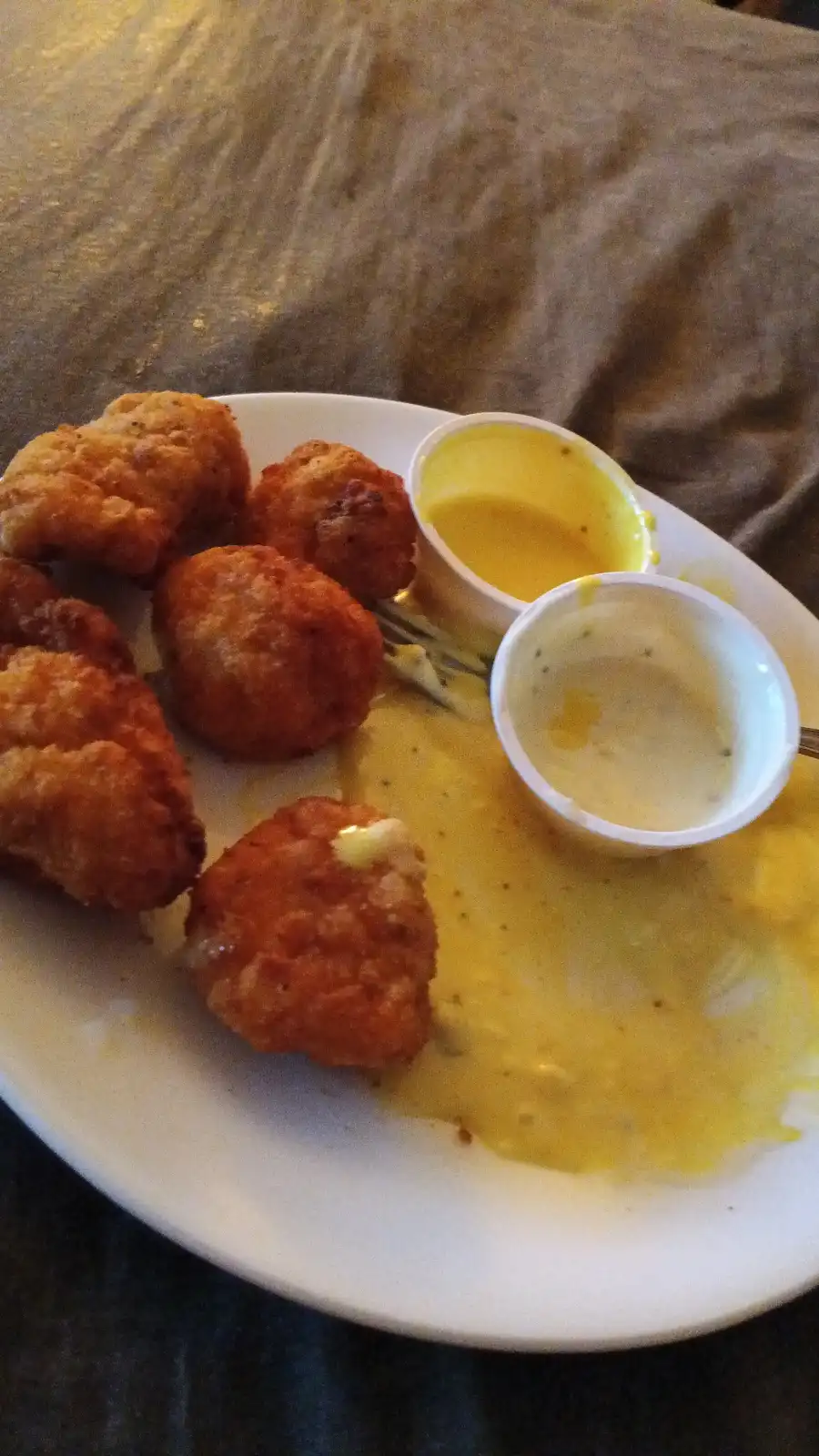 A plate of fried chicken nuggets sits on a table, with a large puddle of yellow sauce spilled across the plate and two small…