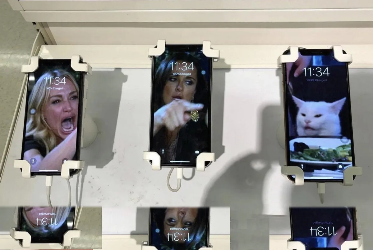 Three smartphones mounted side-by-side in a charging station, each displaying a different popular internet meme image on…