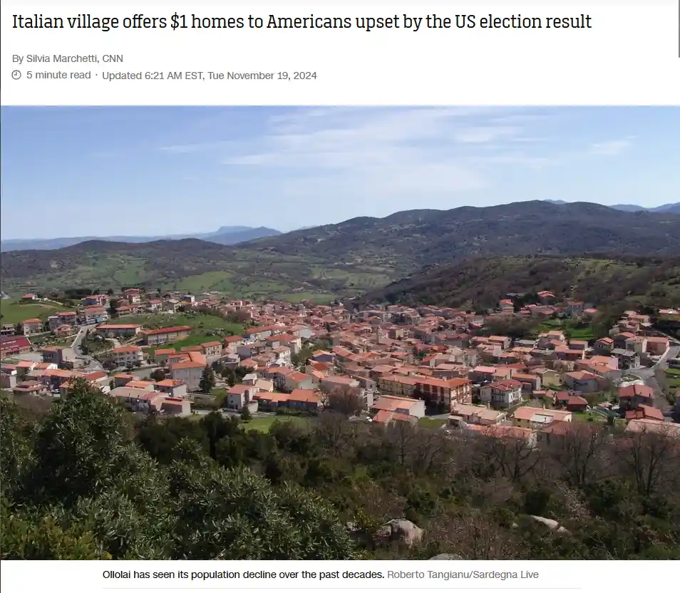 A scenic aerial photograph shows a sprawling Italian village nestled in rolling green hills under a clear blue sky, with…
