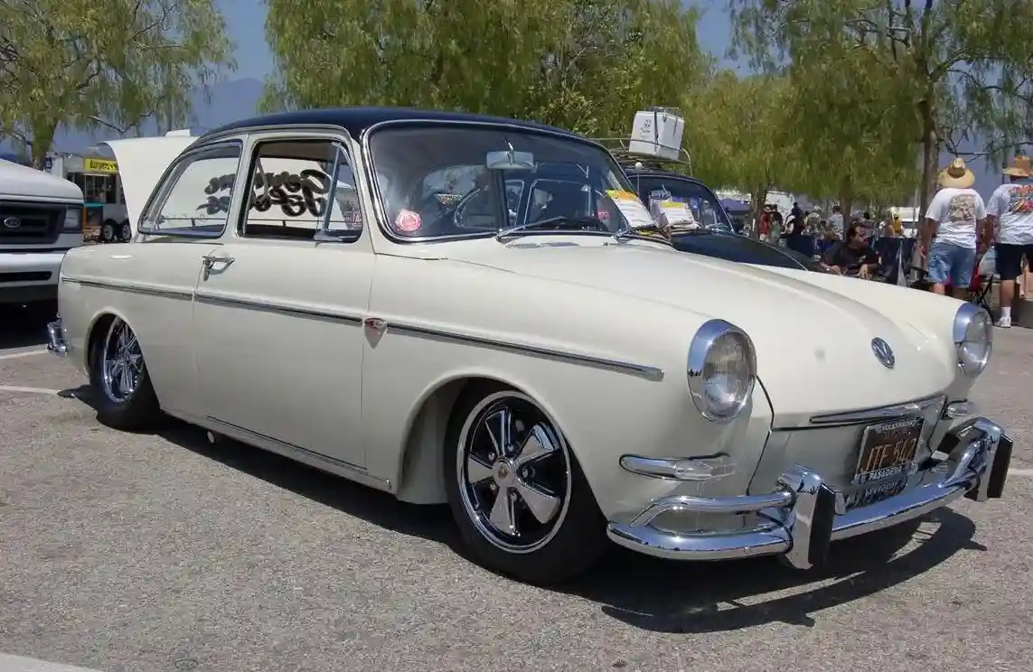 A vintage cream-colored Volkswagen Beetle with chrome bumpers and custom black wheels is parked at an outdoor car show. The…