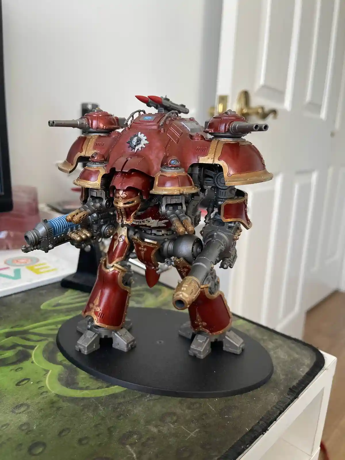 A highly detailed, red and gold painted miniature of a heavily armed, bipedal war machine stands on a black circular base…