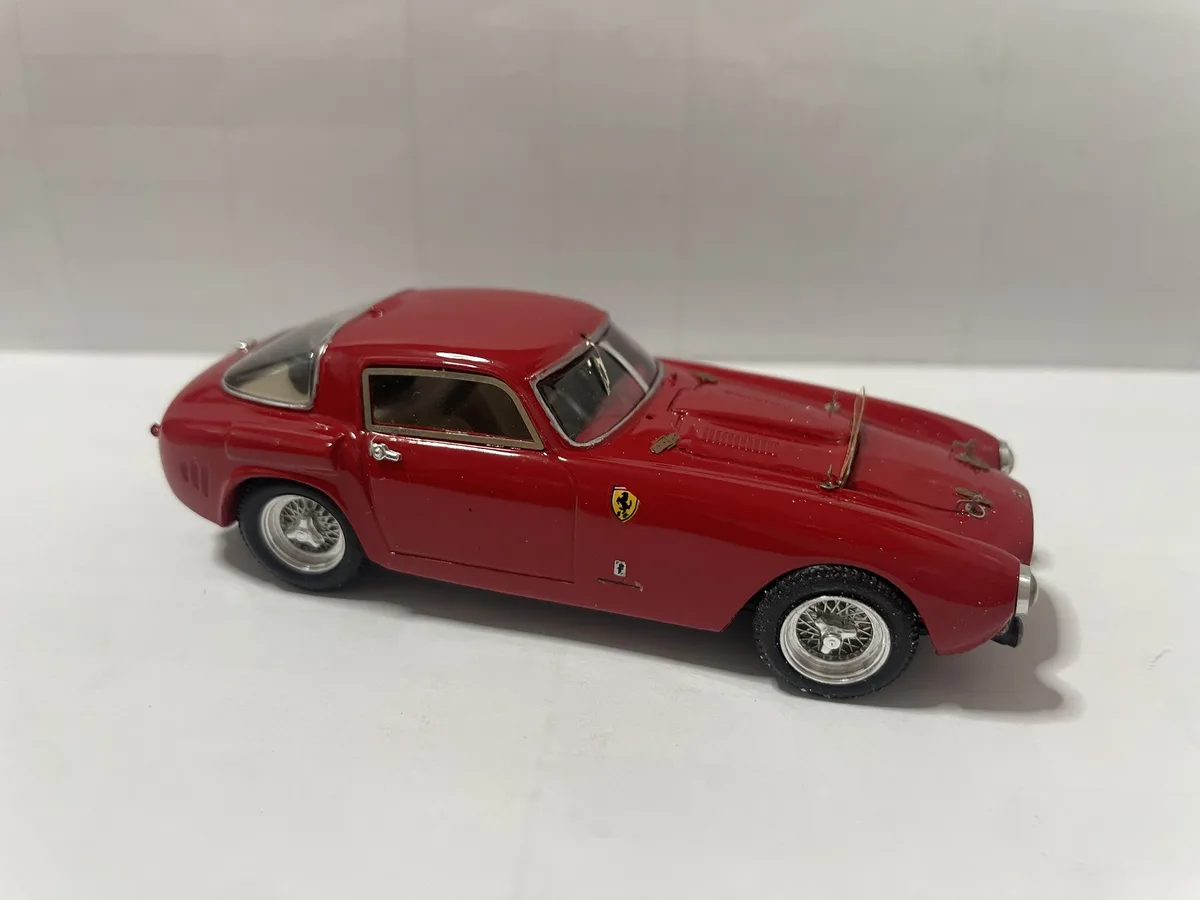 A red die-cast model of a classic Ferrari sports car, shown in profile against a plain white background. The car features a…
