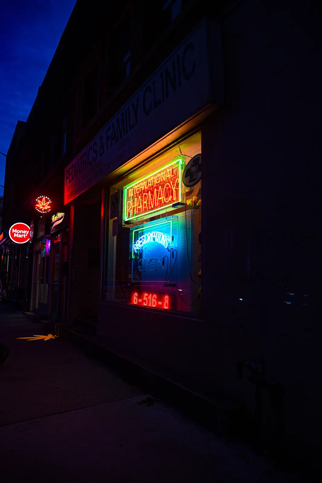 A nighttime street scene shows a brightly lit pharmacy with neon signs reading 'INTERNATIONAL PHARMACY' and 'PRESCRIPTIONS…