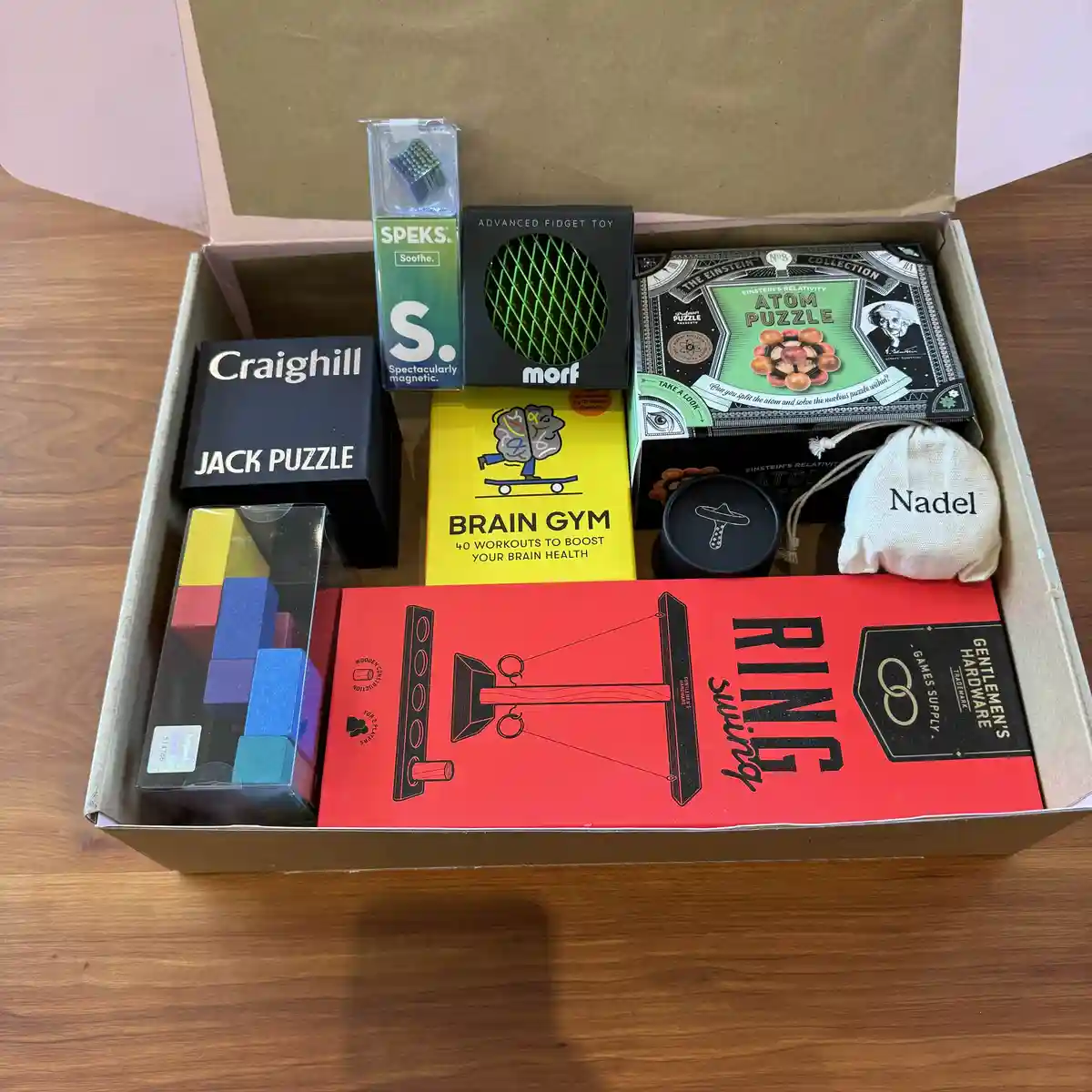 An open cardboard box on a wooden table contains various brain teaser and puzzle products, including a 'Brain Gym' book…