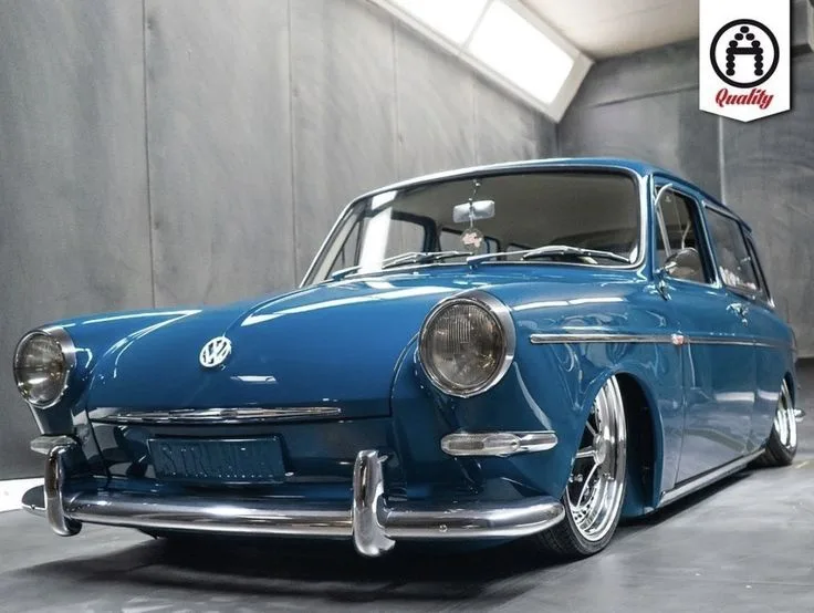 A low-slung, custom-modified blue Volkswagen Type 3 station wagon is photographed in a studio with gray walls and a concrete…
