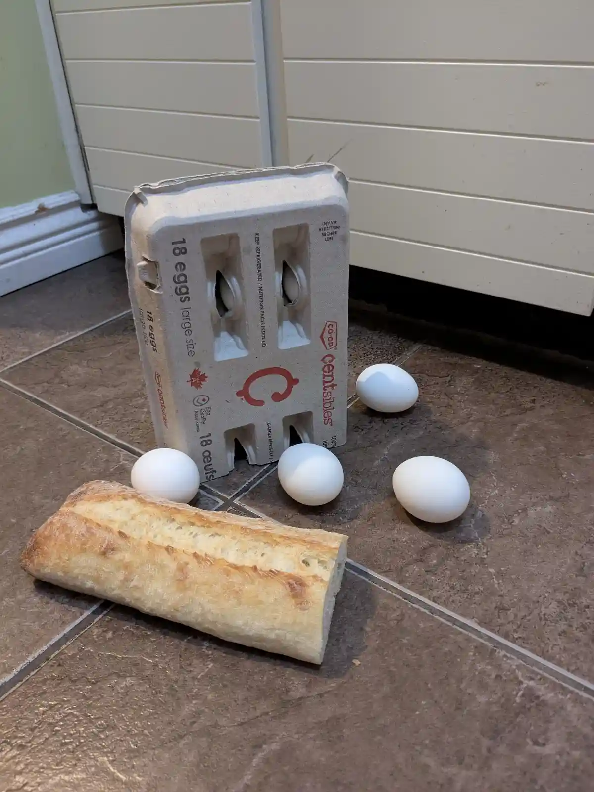 An 18-egg carton from Co-op Centrals stands upright on a tiled floor next to a baguette and four loose white eggs. The…