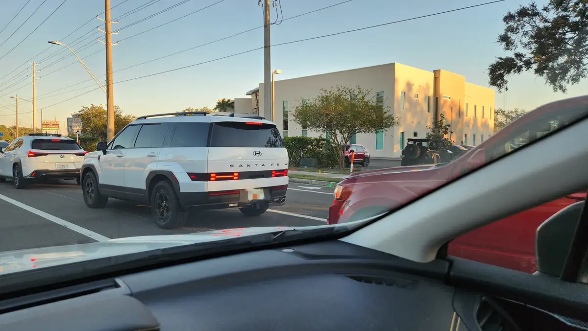 A photo taken from inside a car shows a white Hyundai Santa Fe SUV directly ahead in traffic, with its brake lights on. The…