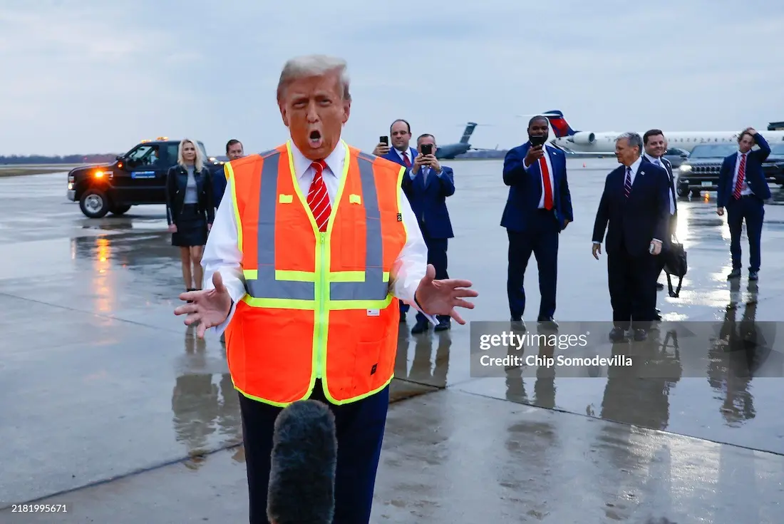 a man in a bright orange safety vest with reflective stripes stands on a wet airport tarmac, mouth open in an exaggerated…