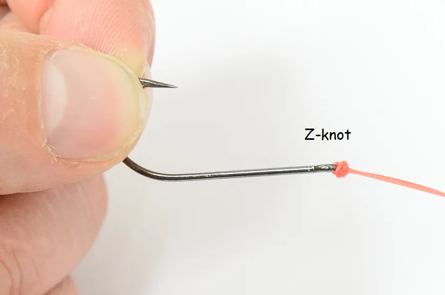 A close-up photo shows a person's thumb and forefinger holding a fishing hook with a bright orange fishing line tied to its…