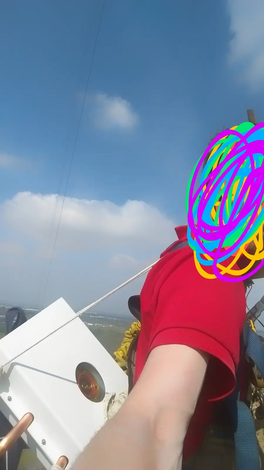 A selfie from a person wearing a red shirt and safety harness, suspended high in the air with a clear blue sky and clouds…