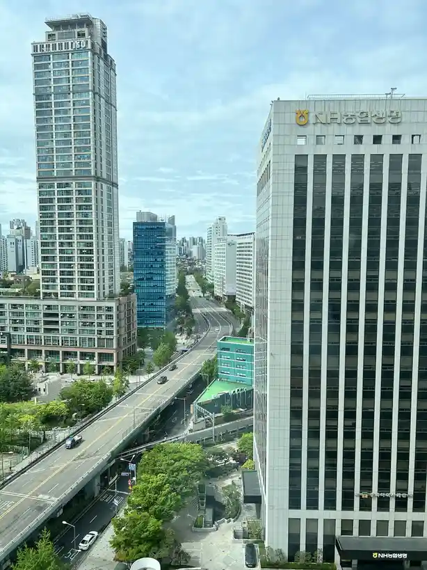 A high-angle cityscape photo shows a tall building with 'NH농협생명' signage on its facade, alongside other skyscrapers and a…