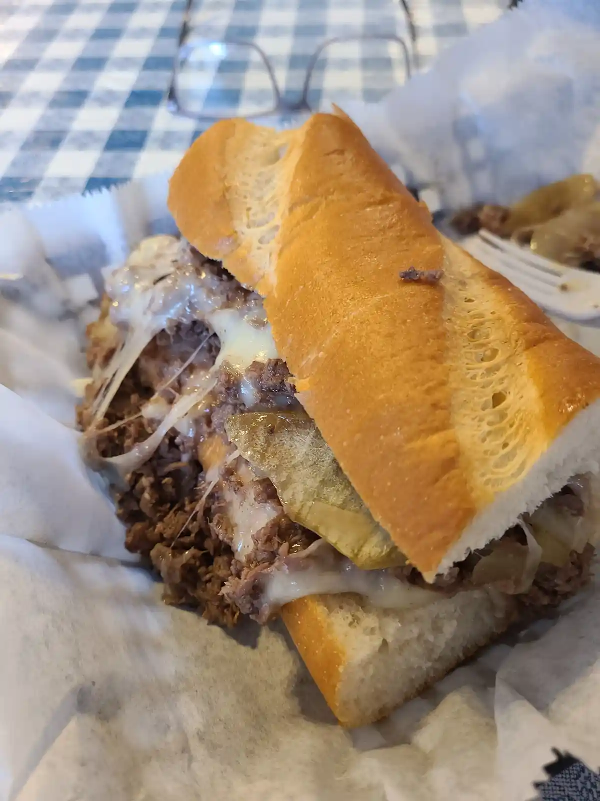 A close-up photo of a cheesy, meat-filled sandwich in a basket lined with paper, resting on a blue and white checkered…