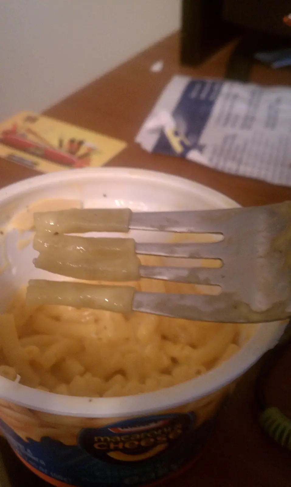 A close-up photo shows a fork lifting three macaroni noodles from a nearly empty Kraft Macaroni & Cheese cup. The noodles…