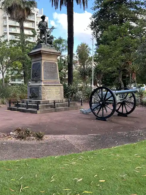 An outdoor photo shows a historical cannon on wheels next to a war memorial statue of soldiers, set in a park with palm…