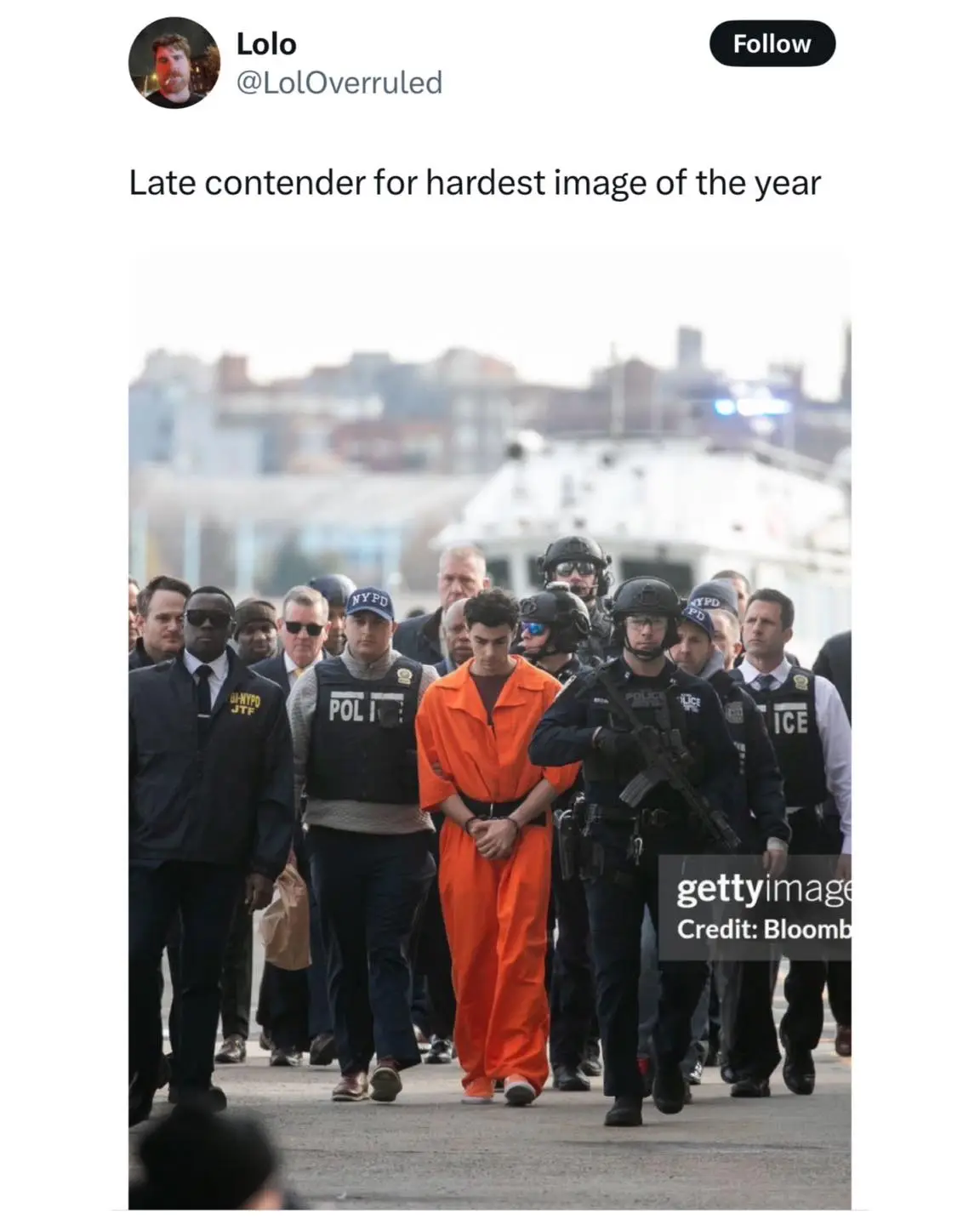 A young man in an orange prison jumpsuit is being escorted by multiple heavily armed NYPD officers through a crowd. The…
