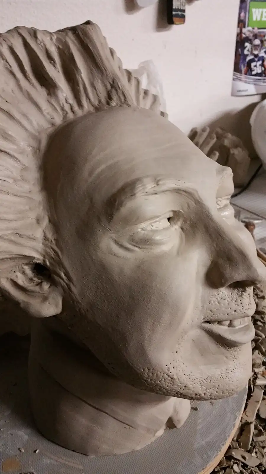 A close-up of an unfinished clay sculpture of a man's head, showing detailed facial features and a spiky, textured…