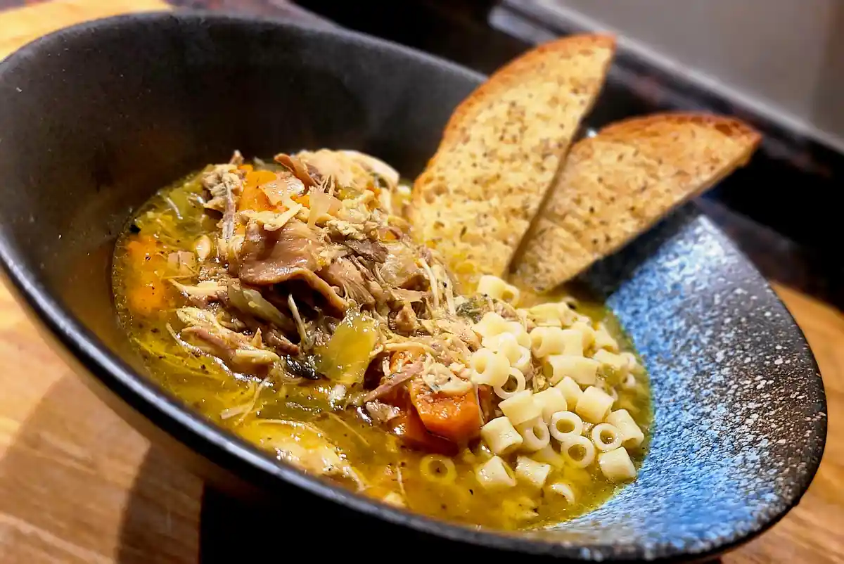 A close-up photo shows a dark ceramic bowl filled with a hearty, golden-brown soup containing shredded meat, carrots, and…