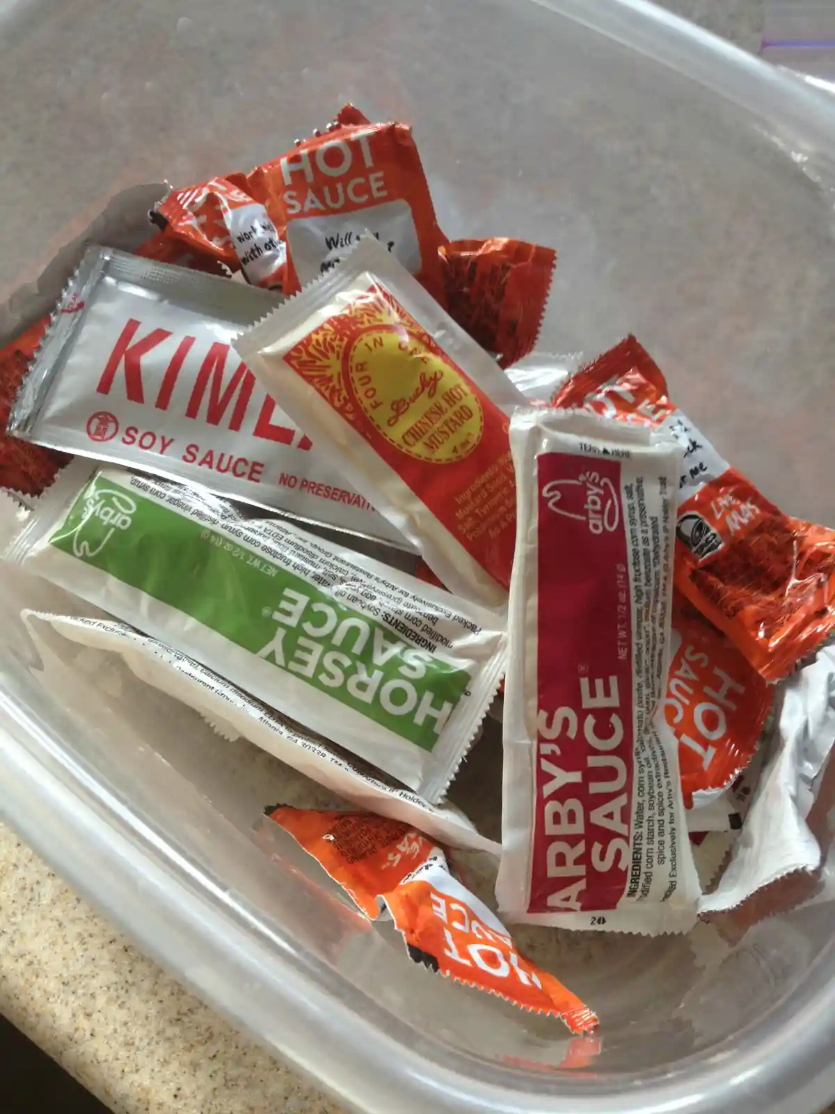 A clear plastic container holds a jumble of various restaurant sauce packets, including Arby's sauce, Kimchi soy sauce…