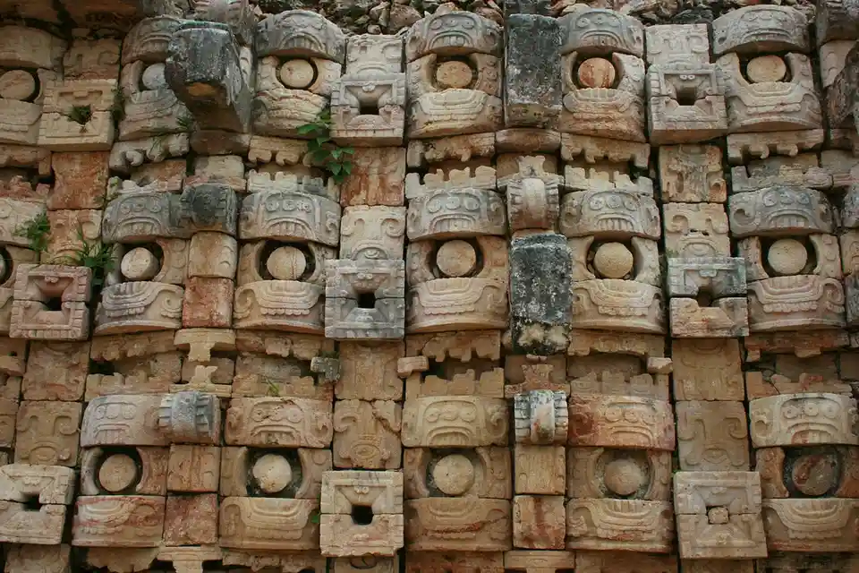 a close-up photograph of a weathered stone wall composed of numerous carved, rectangular blocks featuring stylized Mayan or…