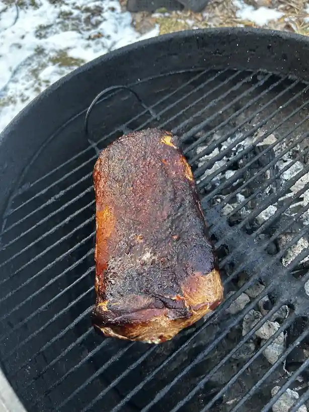 A large, rectangular piece of meat with a charred, seasoned crust sits on the grates of a black charcoal grill, with gray…