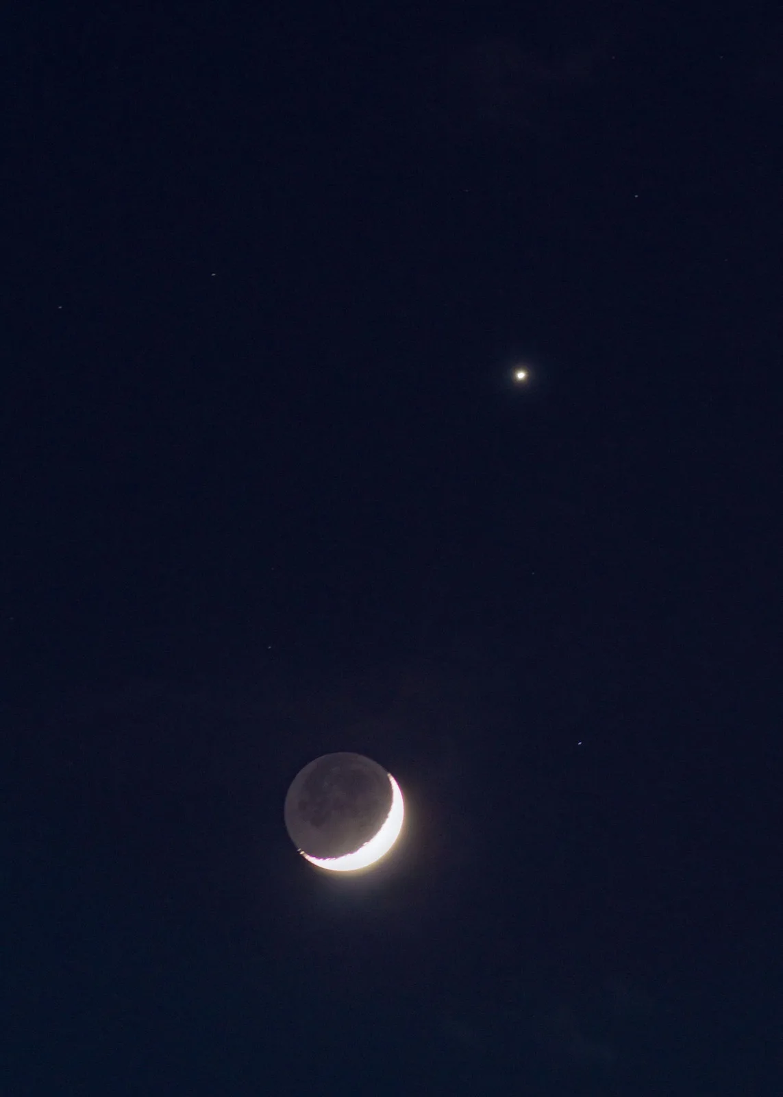 A crescent moon is visible in a dark night sky, with a bright star or planet positioned above it. The moon's illuminated…