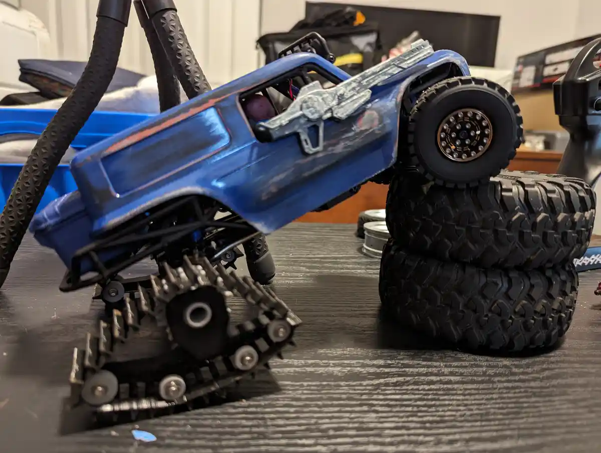 A blue remote-controlled monster truck with a tank tread on its front is balanced precariously on a stack of two black…