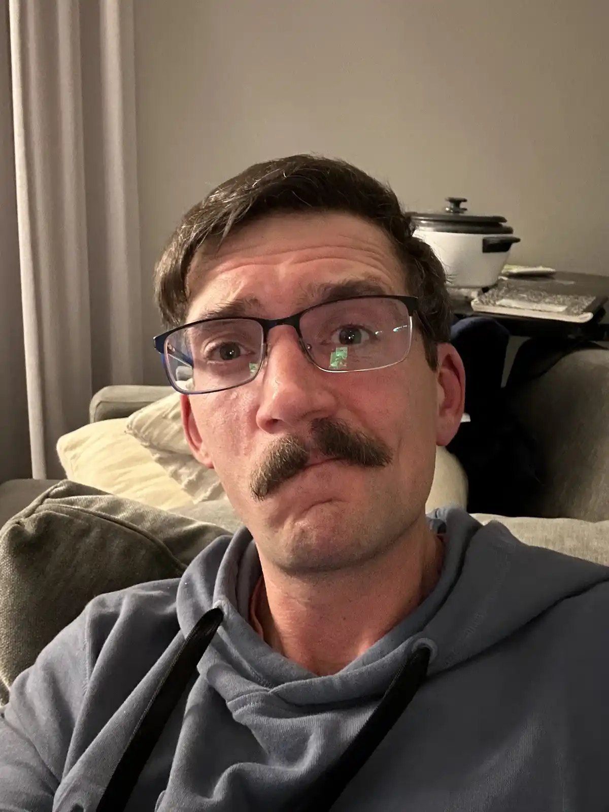 A man with glasses and a mustache sits on a couch, making a tired, unimpressed face. Behind him, a slow cooker and laptop…