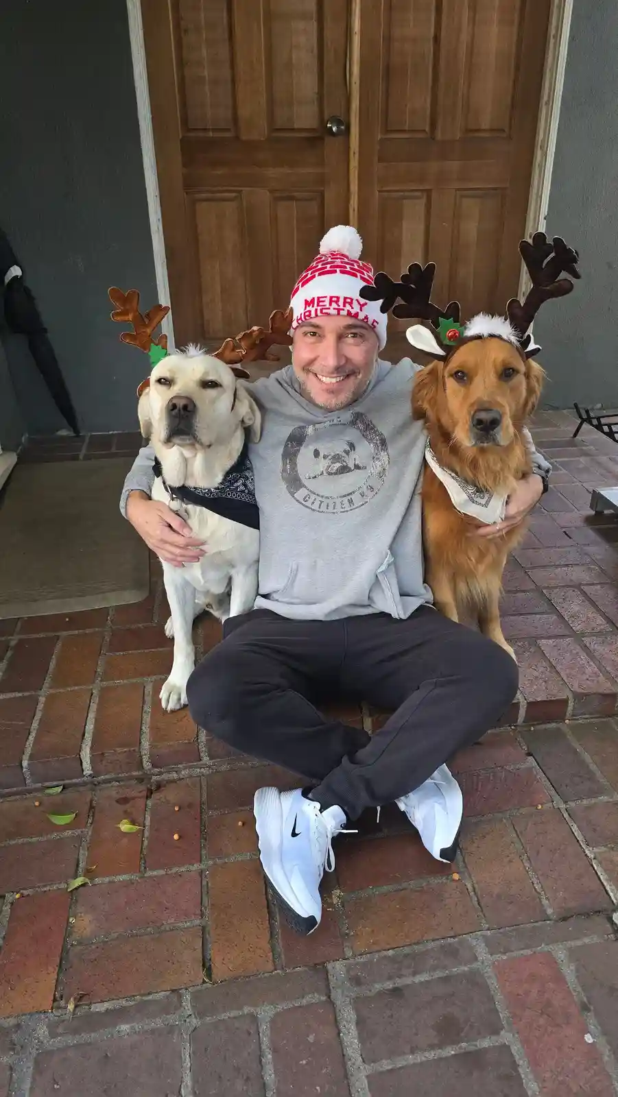 A man wearing a 'merry christmas' hat sits on a brick porch with two dogs, each wearing reindeer antlers. The man is smiling…