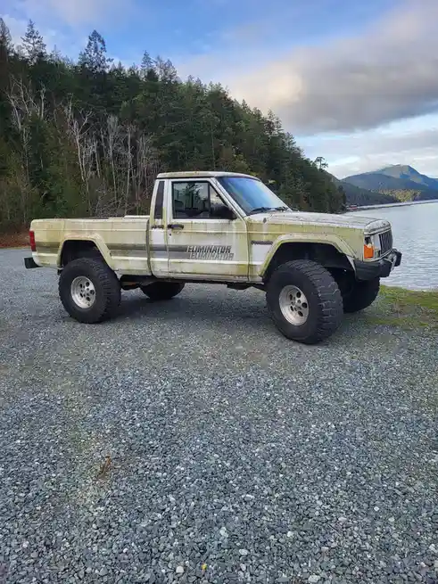 A heavily modified, rusty, and dirty white Jeep Comanche pickup truck with 'ELIMINATOR' written on its side is parked on a…