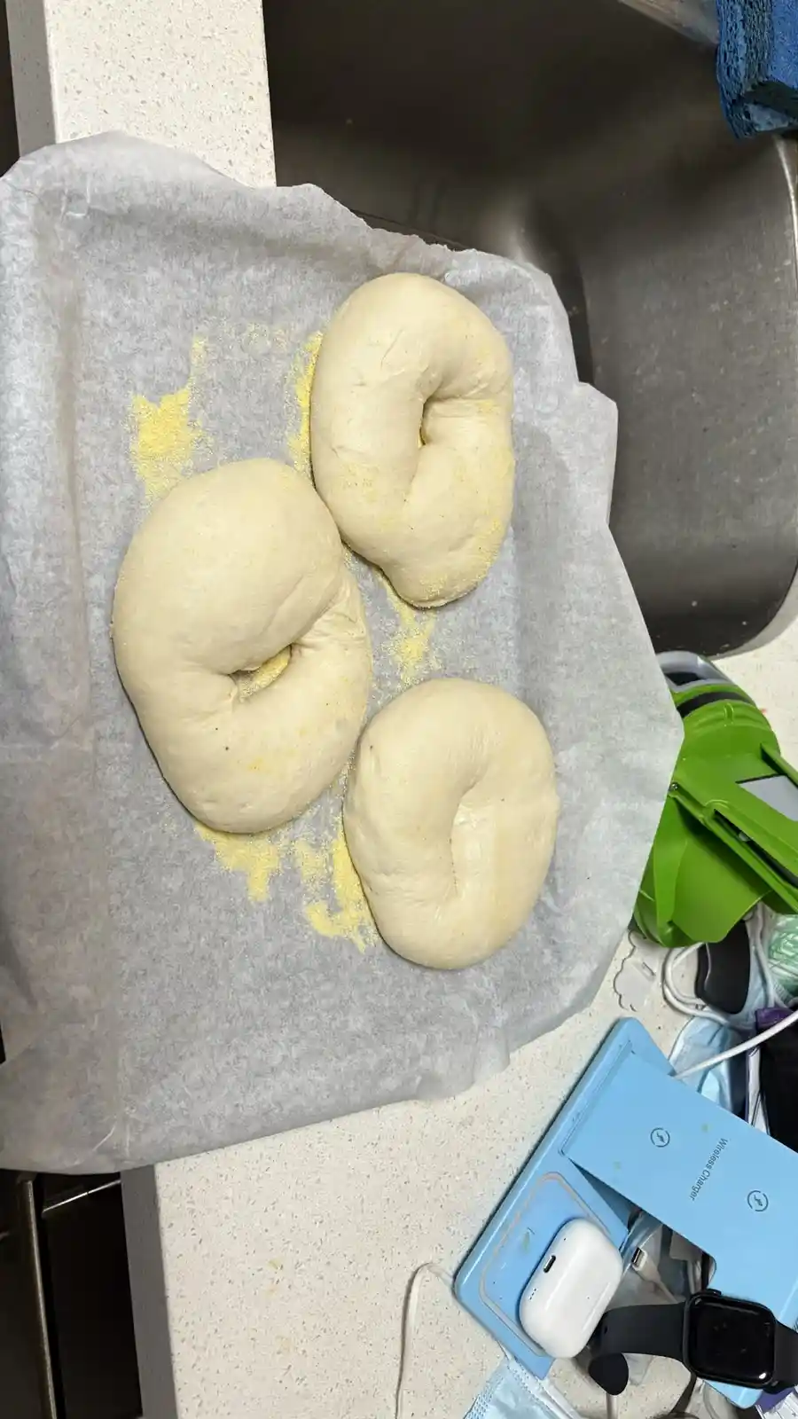 Three raw bagels sit on parchment paper next to a sink, with yellow flour dusting around them. The overlaid text reads 'i…
