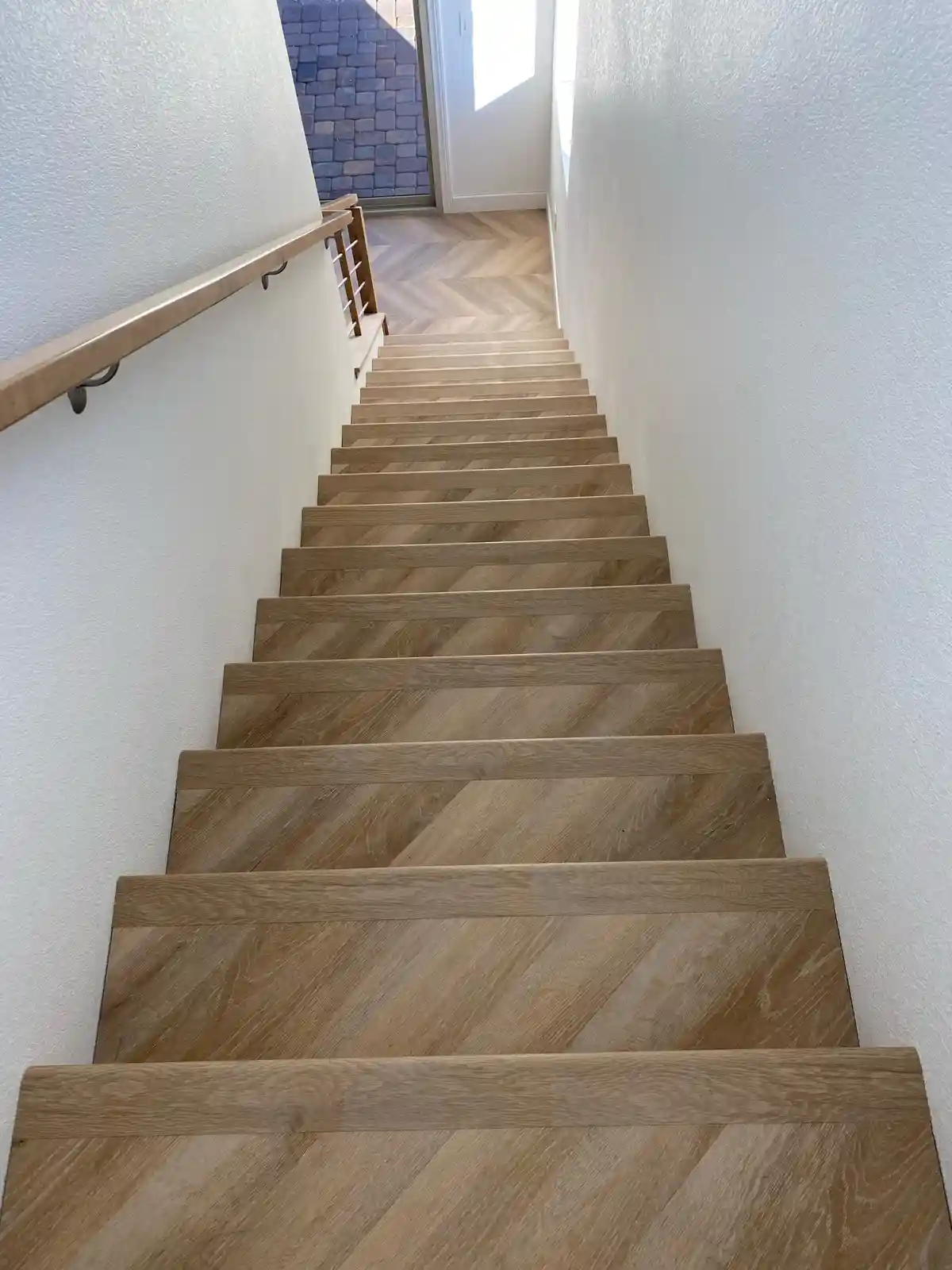A low-angle photo looking up a long, narrow staircase with light wood treads and white walls, ending at a doorway with a…