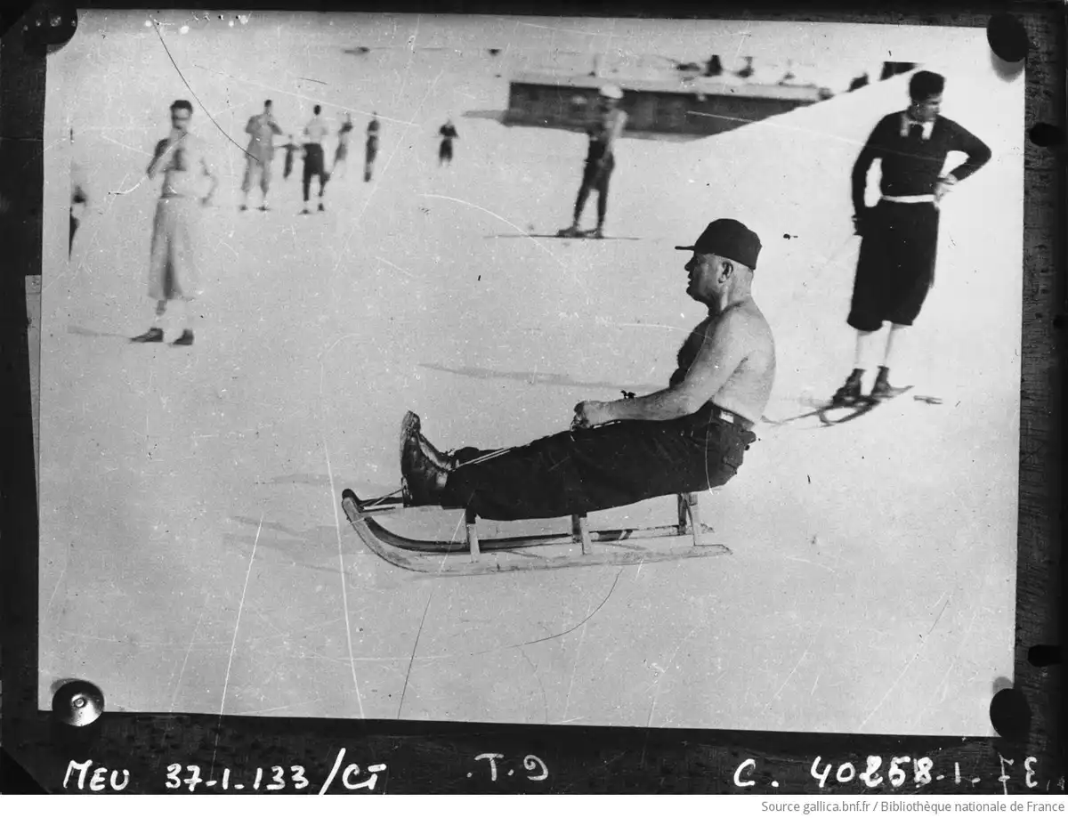 a black-and-white historical photo shows a shirtless man sitting on a sled in the snow, looking off to the side while other…